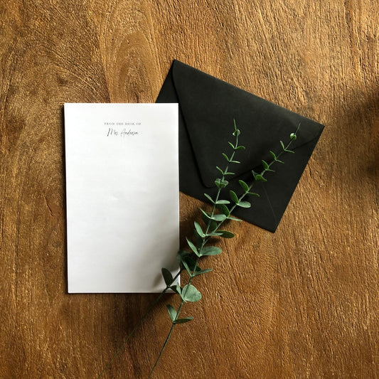 White personalized notepad with black envelope and green branch on wooden surface