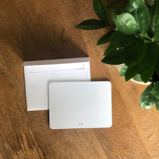 White monogrammed notecard on a wooden surface with a plant in the corner.