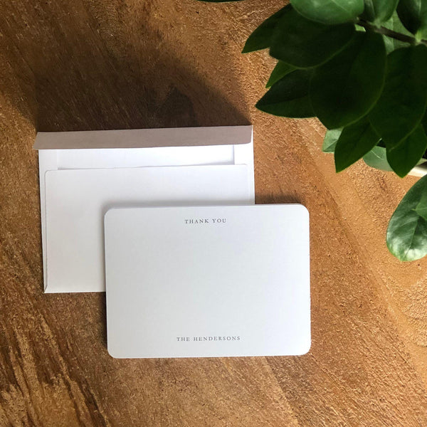 White note card with 'Thank You' text on a wooden surface with a plant in the corner