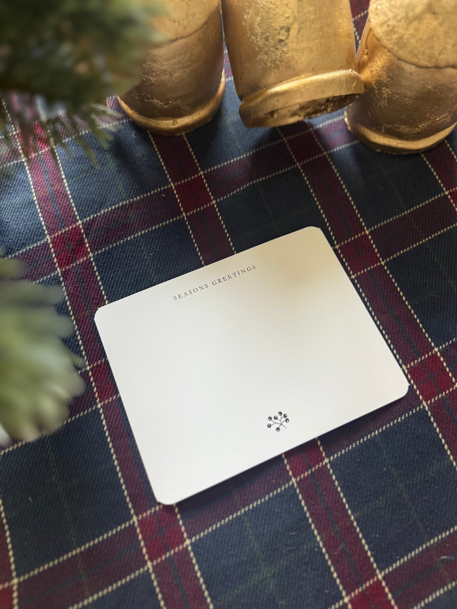 White Seasons Greeting note card with text on a plaid tablecloth with gold bells